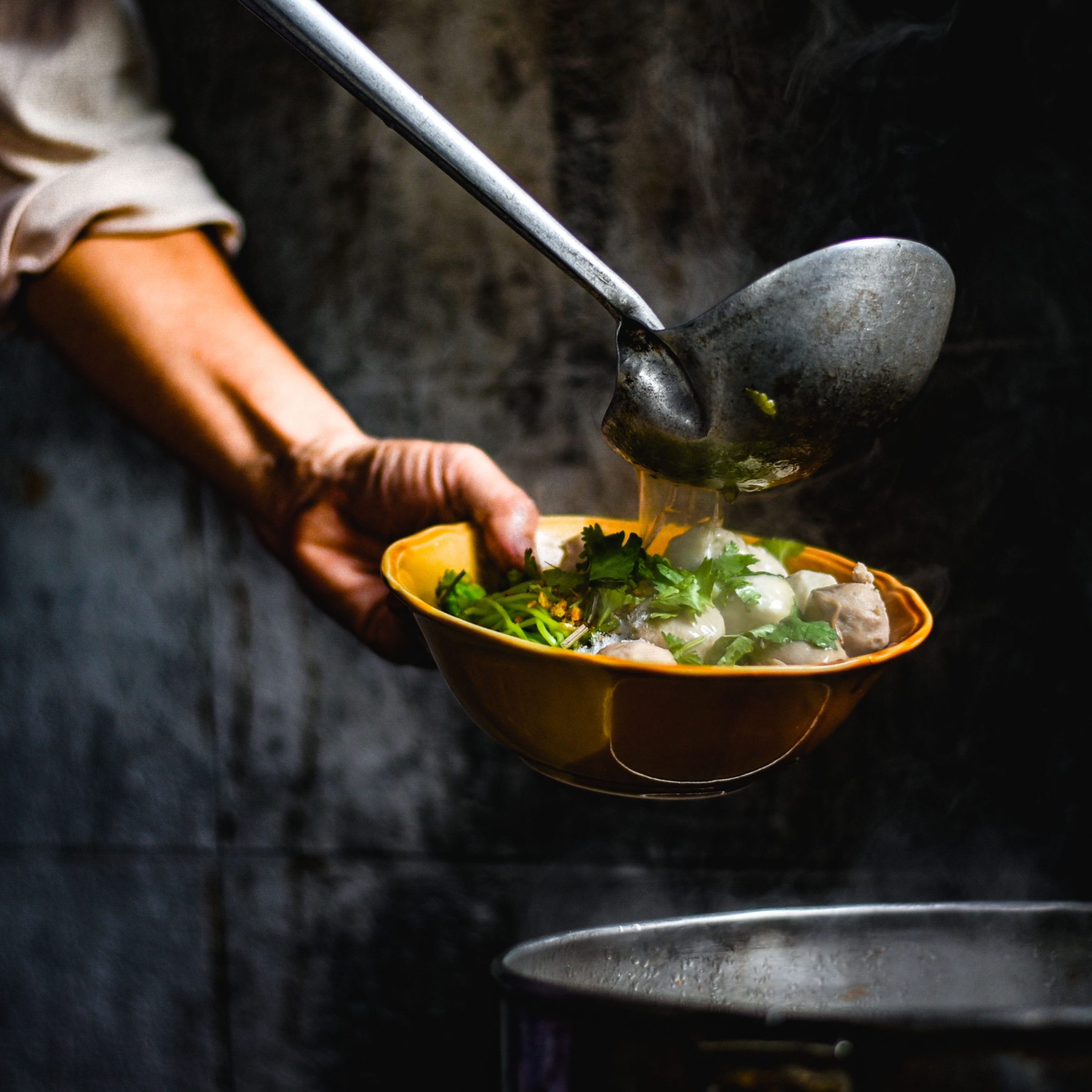 Hands of Thai Chef are scooping a delicious soup topped with noodles and pork balls in the corner of the restaurant with light shining to the noodle bowl that is traditional Thai food.